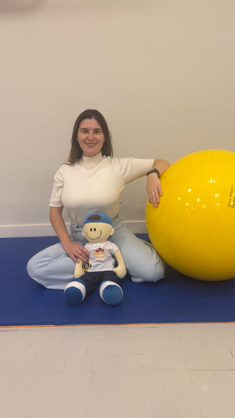 Dra. Giovanna Luvizotto sorrindo ao lado de um boneco de fisioterapia sobre uma bola suíça amarela, em consultório de atendimento infantil.